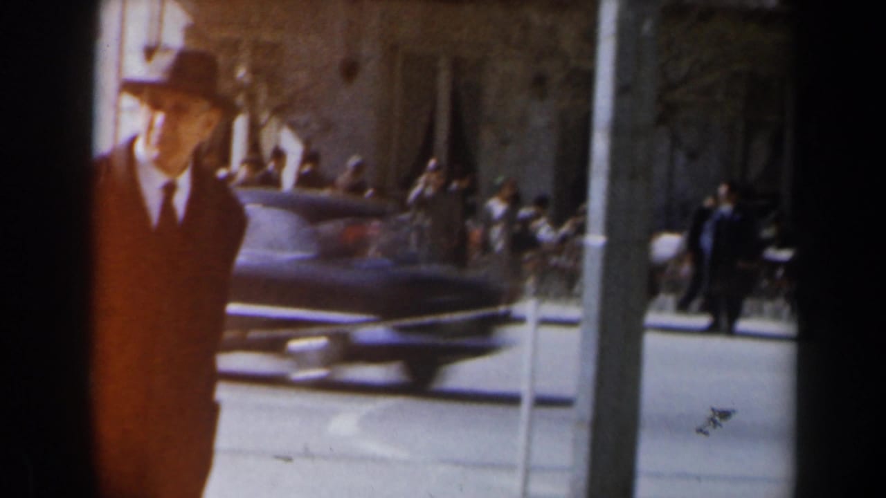 A man in a hat stands or walks near the curb as a blue car passes behind him.