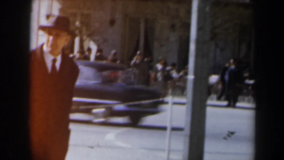 Street-level 1960s Israel view of a suited man in a brimmed hat near passing traffic and pedestrians.