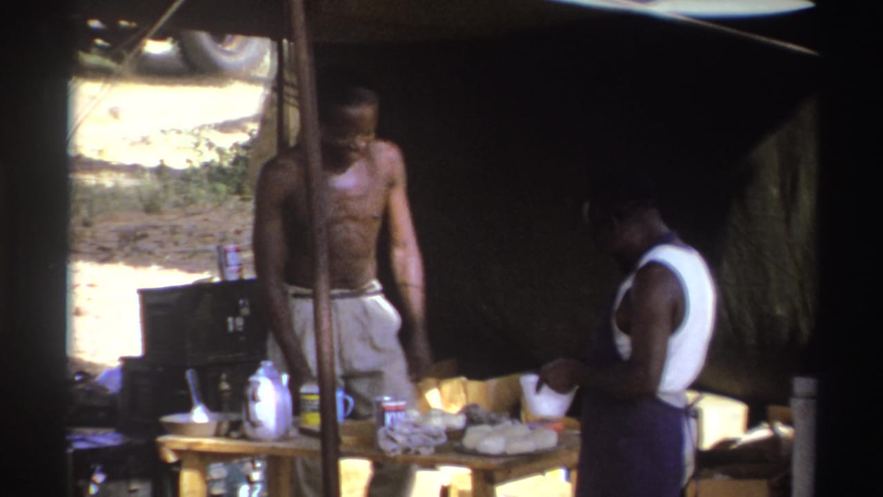 In 1969 South Africa home-movie footage, two men work under a makeshift tent beside a table holding containers and food items.