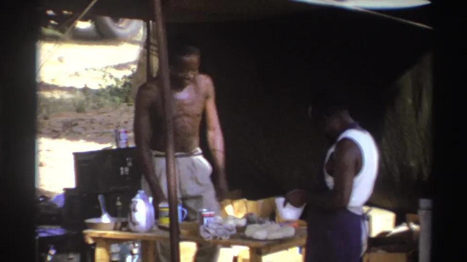 In 1969 South Africa home-movie footage, two men work under a makeshift tent beside a table holding containers and food items.