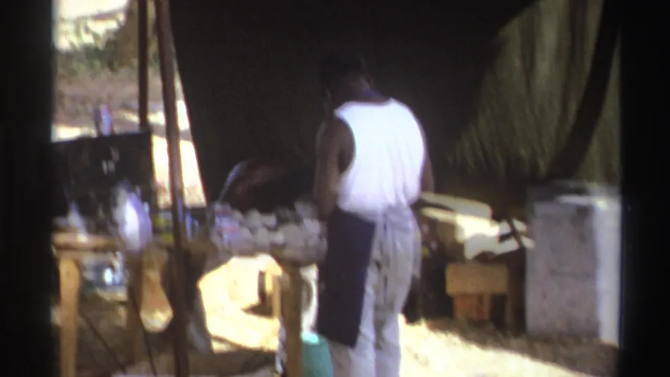 A man in a white tank top and apron stands at a wooden table in a shaded South African outdoor cooking setup from 1969 footage.