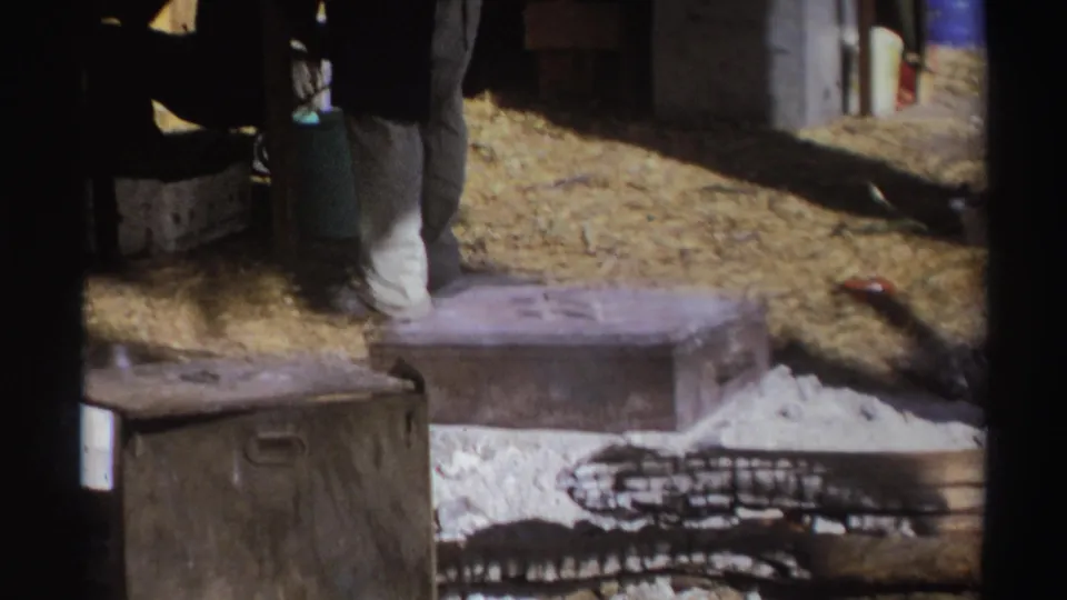 South Africa 1969 home-movie frame showing a person near supplies, metal chests, straw-covered ground, firewood, and a low fire area.