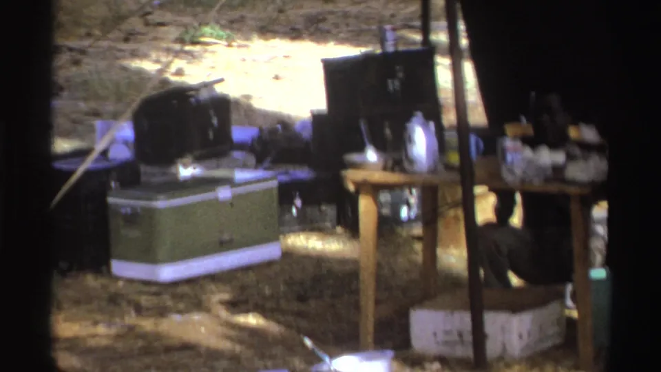 1969 South Africa 8mm frame showing camping supplies, a green cooler, table, kettle, and partly visible adults in a tented kitchen setup.