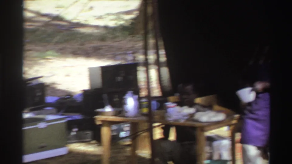1969 South Africa campsite frame showing a tent kitchen with cooler, table, utensils, dark hanging fabric, and a partly visible adult.