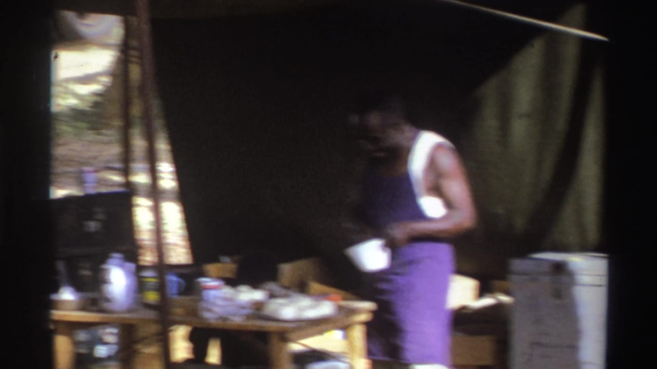 1969 South Africa 8mm frame showing a man in a purple apron preparing food at a wooden table inside a makeshift tent.