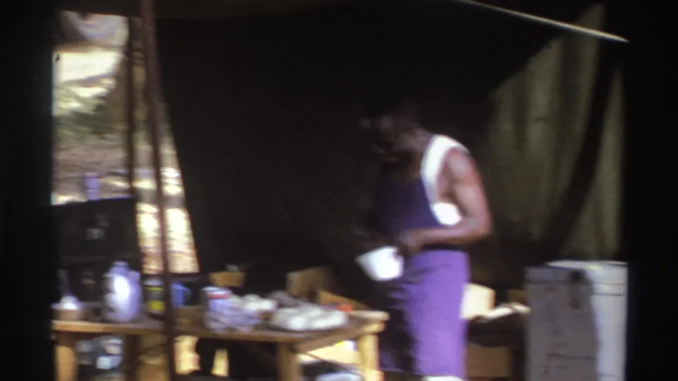 1969 South Africa 8mm frame showing a man in a purple apron preparing food at a wooden table inside a makeshift tent.