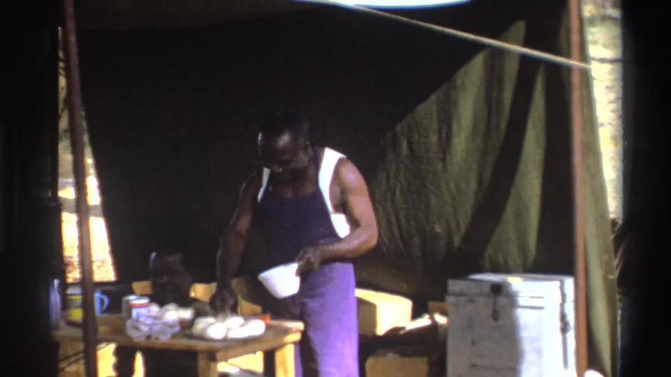 1969 South Africa home-movie frame with one man holding a bowl at a camp table and another man seated in the tent.