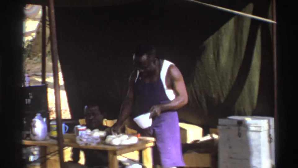 8mm frame from 1969 South Africa showing a man preparing food on a small table inside a tent while another man sits close by.