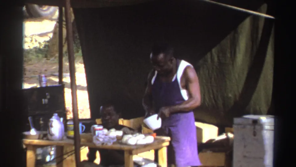 1969 South Africa 8mm frame showing a man in a purple apron handling a bowl at a camp table in a shaded tent.