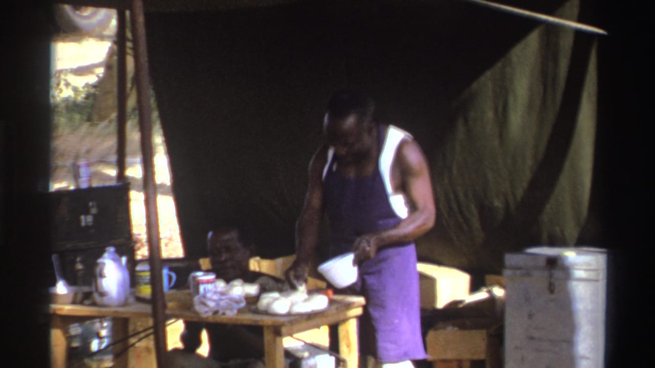 In 1969 South Africa home-movie footage, a man in a purple apron handles a bowl at an outdoor table under a tarp.