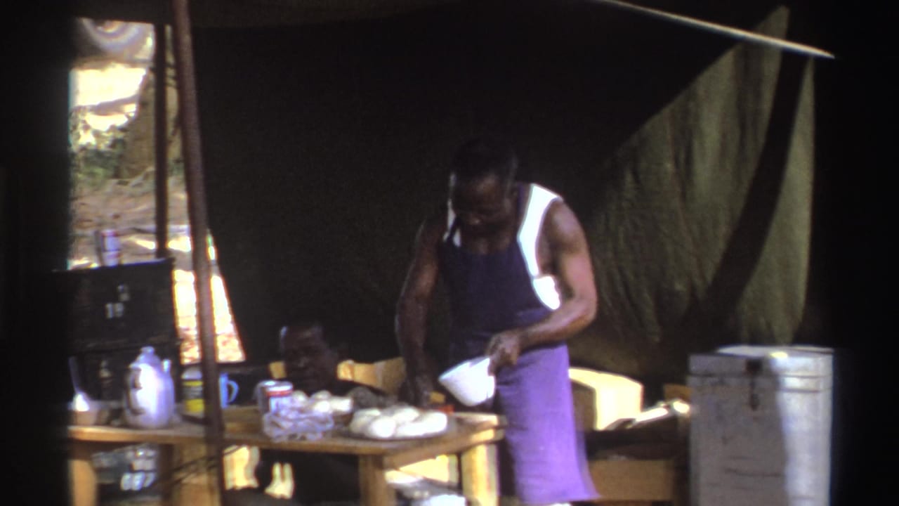 Man leans over a table with a bowl and food items beneath a tarp.