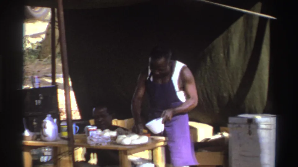 A man in a purple apron leans over an outdoor food table in South Africa, with a kettle and supplies beside him.