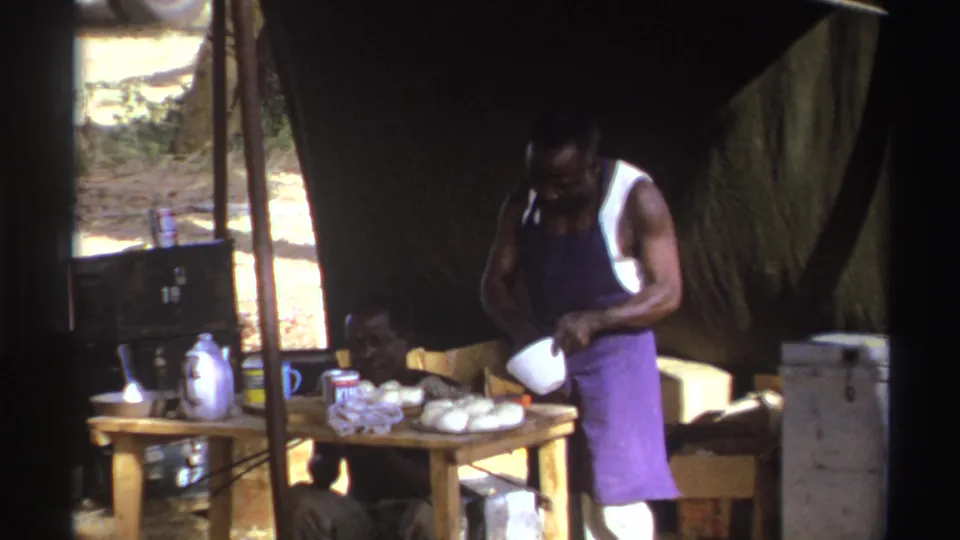 The 8mm frame shows a man holding a white bowl beside bread or food items at a shaded outdoor station.