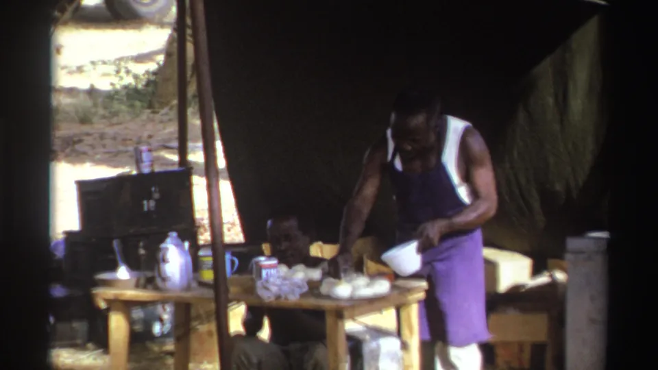 A man in a purple apron bends toward a wooden food table under a tarp, with daylight visible beyond the shelter.