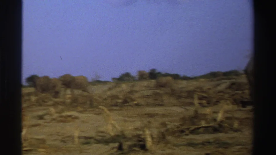 1969 South Africa 8mm frame showing dry cleared ground, sparse brush and a distant elephant under a bright blue sky.