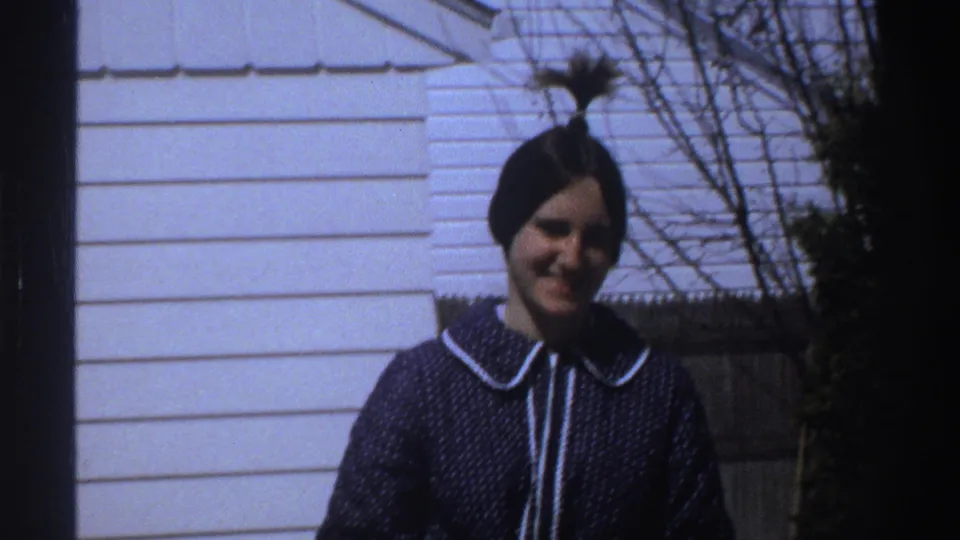 A woman with dark hair tied up smiles near a white-sided house and fence in 1969 home-movie footage from South Africa.
