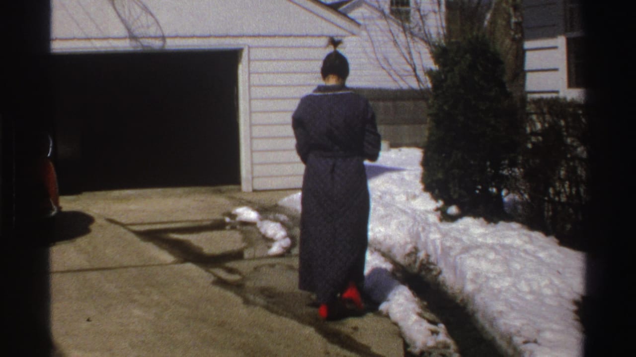 In a 1969 Michigan home-movie frame, a woman in a dark robe stands with her back to the camera near an open garage and snow-lined driveway.