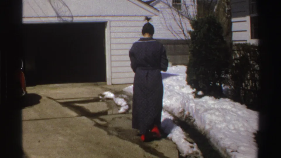 In a 1969 Michigan home-movie frame, a woman in a dark robe stands with her back to the camera near an open garage and snow-lined driveway.