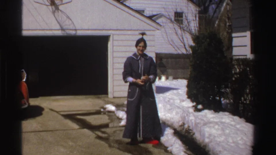 In a 1969 Michigan driveway scene, the woman turns toward the camera in a dark robe with red slippers, standing by an open garage and snow.
