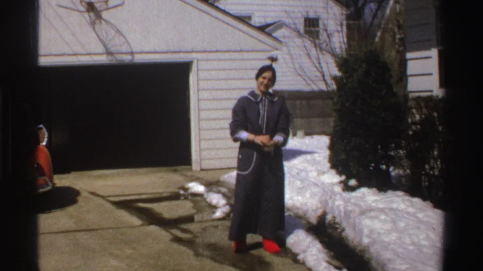 The 1969 Michigan home-movie frame shows a smiling woman in a dark robe and red slippers standing on a sunny driveway beside snow.