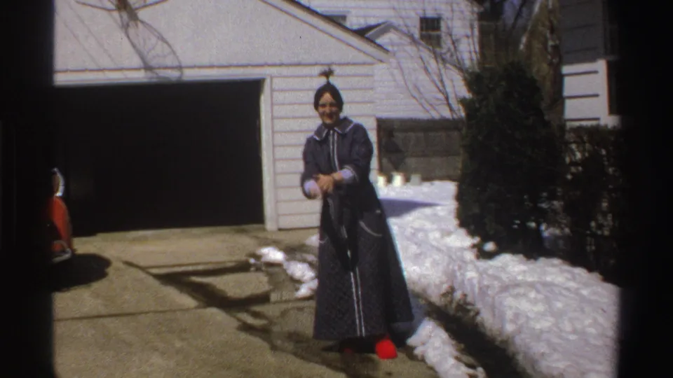 A woman in a dark robe and red slippers smiles toward the camera near an open garage in this 1969 Michigan winter exterior.