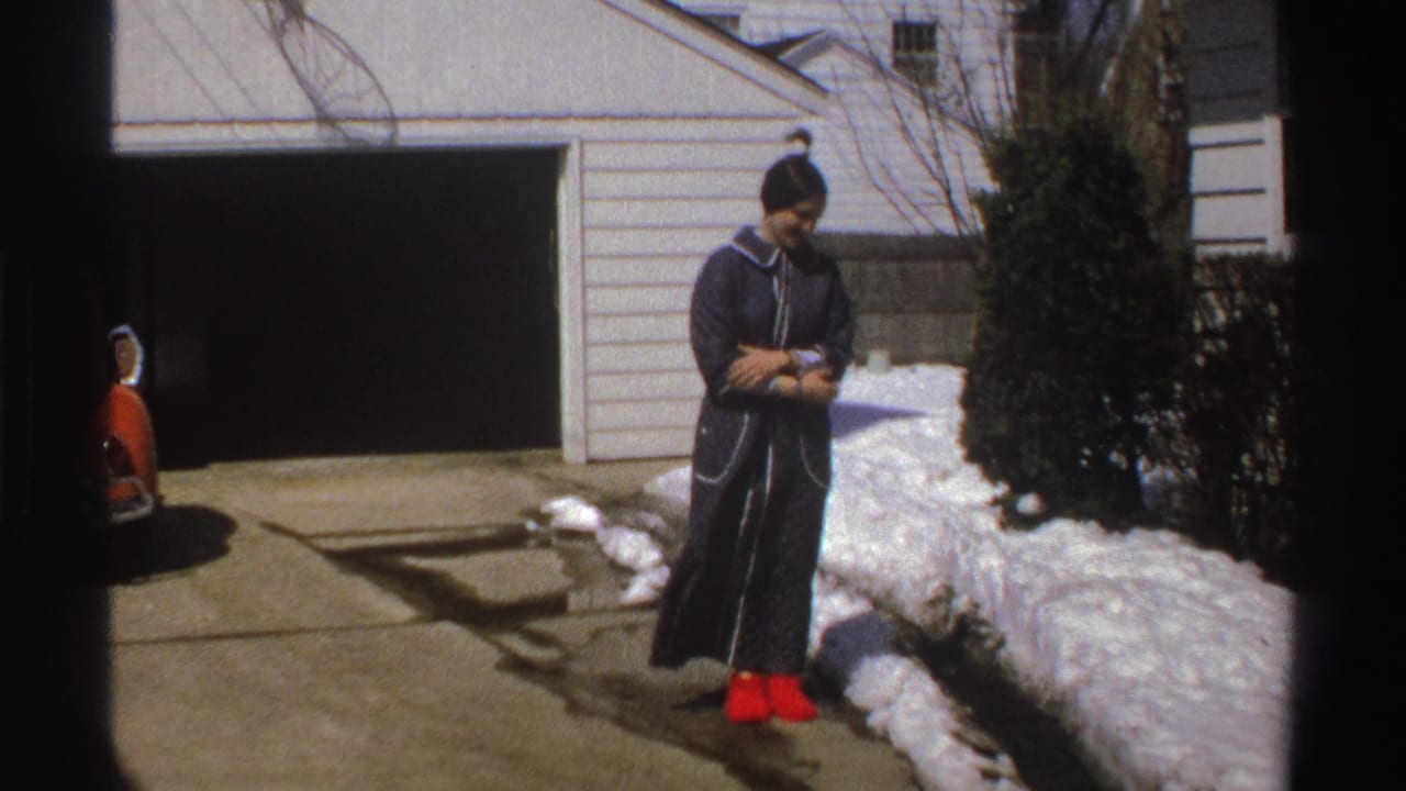 In 1969 Michigan home-movie footage, a woman in a dark coat and red boots stands near snow in a suburban driveway.