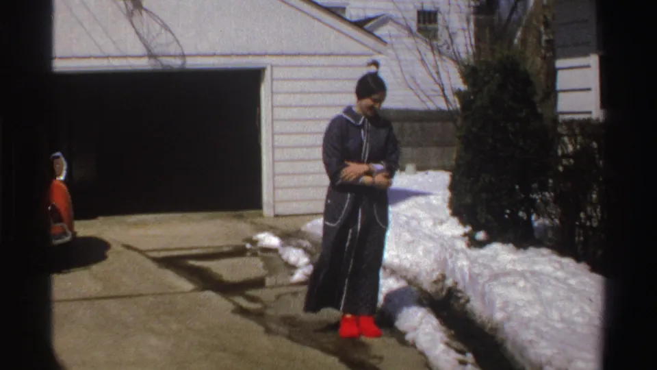 In 1969 Michigan home-movie footage, a woman in a dark coat and red boots stands near snow in a suburban driveway.