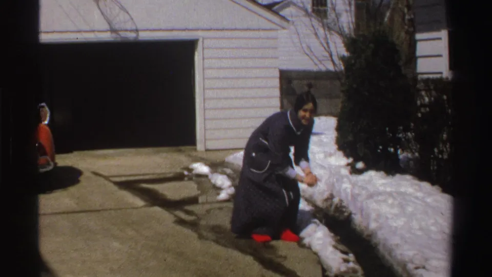 The woman bends down toward snow along the driveway, with a garage, shrubs, and house siding behind her.