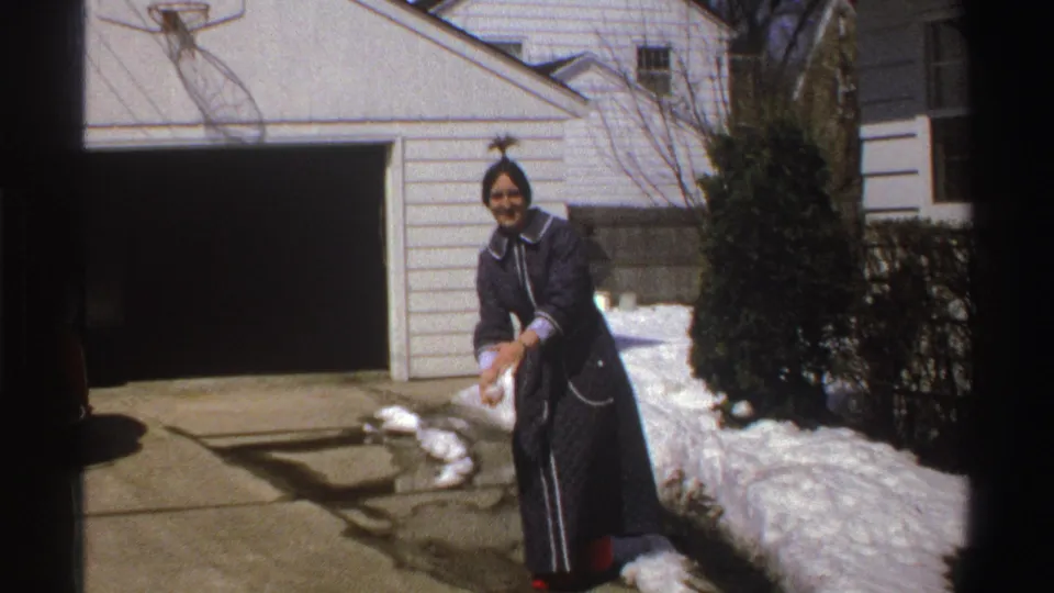 The woman in a dark coat smiles as she shapes a snowball near the driveway snowbank outside a suburban house.