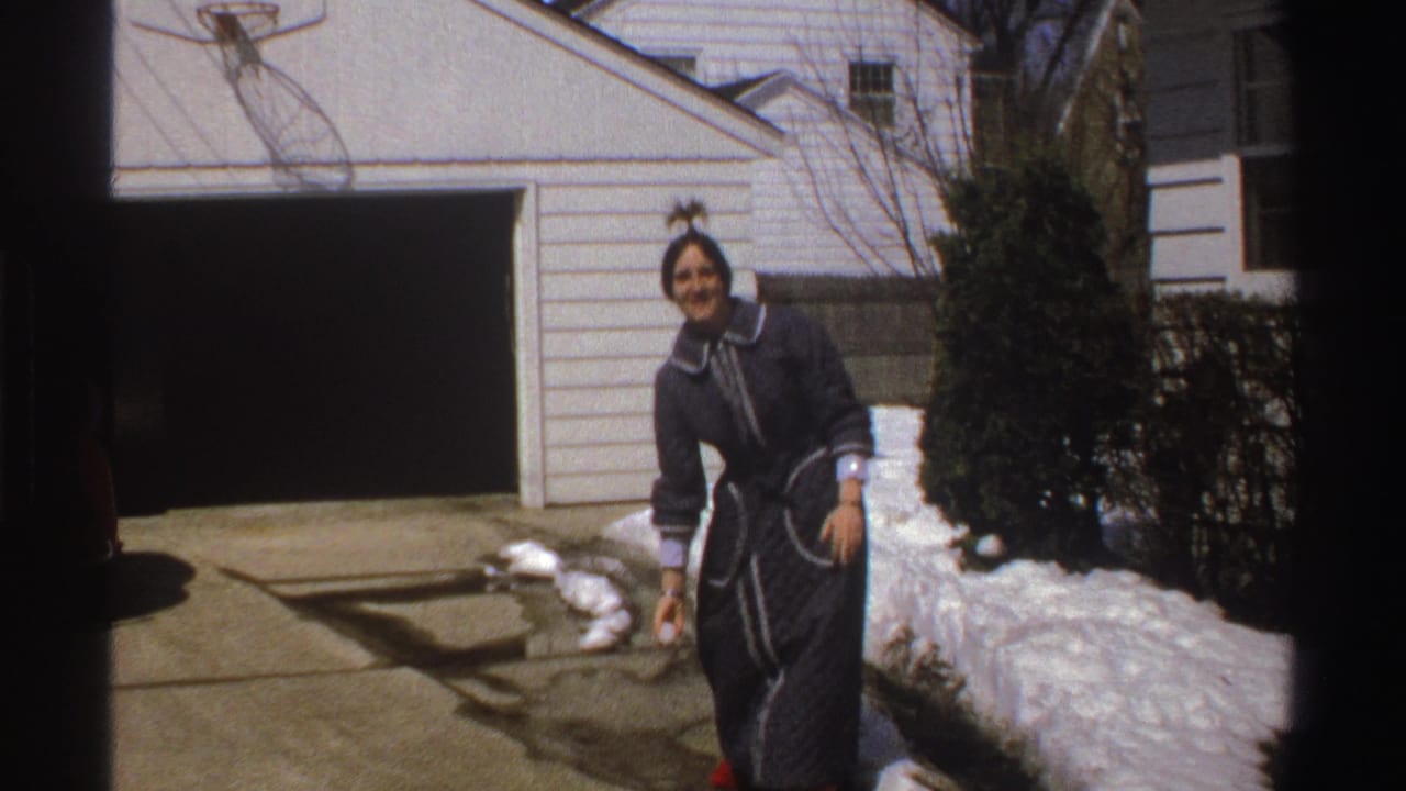 In Michigan home-movie footage from 1969, a woman in a dark robe and red slippers smiles in a snowy driveway.