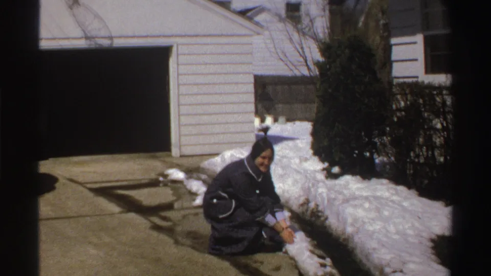 The woman crouches at the edge of a snowy Michigan driveway, appearing to gather snow in 1969 home-movie footage.