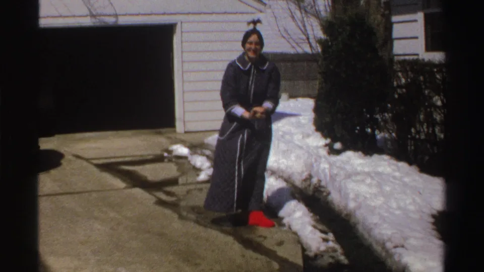 A woman in a dark robe stands in the snowy driveway, holding snow while facing the camera in Michigan, 1969.