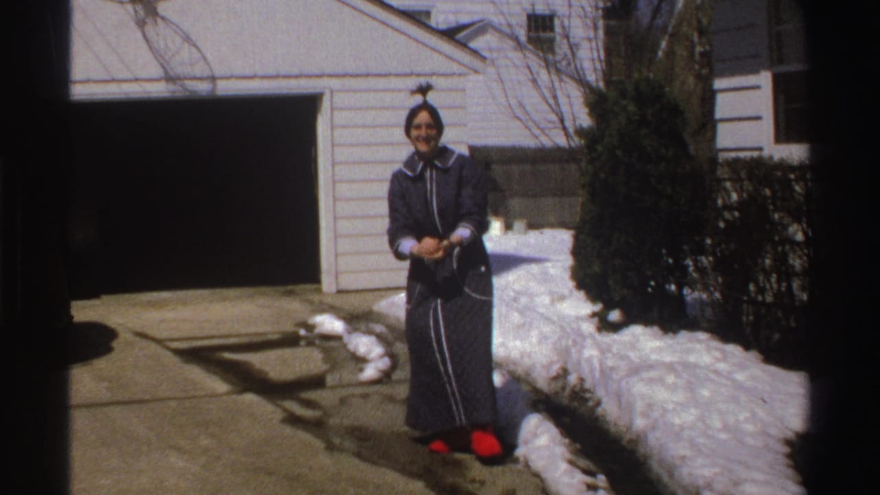 In Michigan home-movie footage from 1969, a woman in a dark robe and red slippers stands outside by snowbanks and a two-car garage.