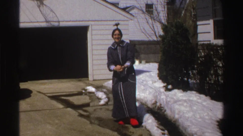 In Michigan home-movie footage from 1969, a woman in a dark robe and red slippers stands outside by snowbanks and a two-car garage.