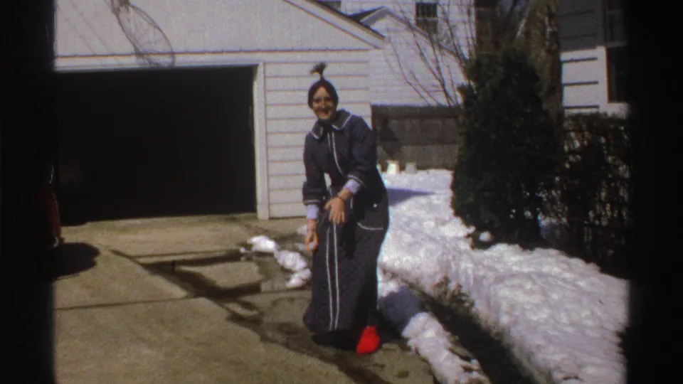 The woman appears to prepare or gather snow in a Michigan driveway, with a garage, shrubs, and snowbanks behind her.