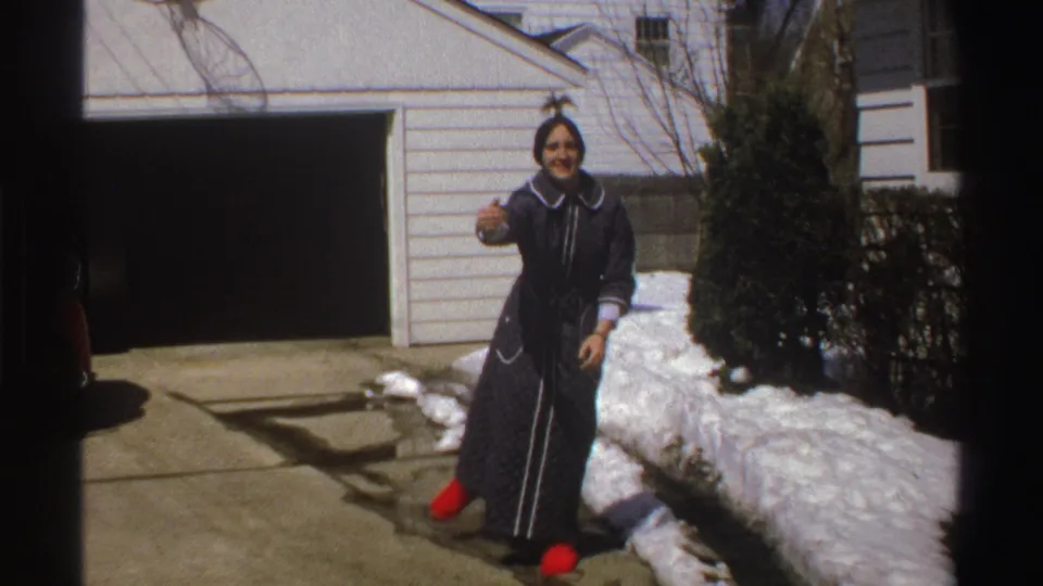 A woman in red slippers throws a snowball toward the camera in a snowy Michigan driveway in 1969 home-movie footage.