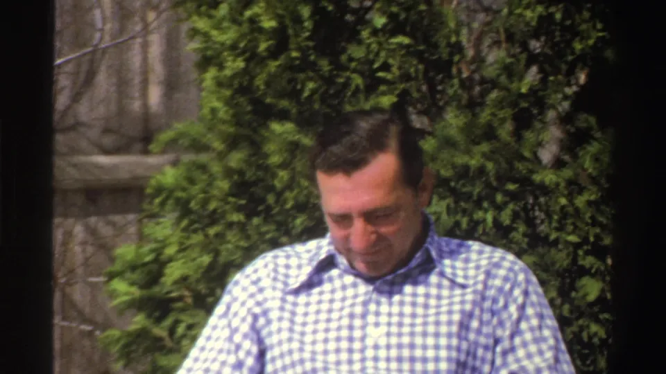 A man in a blue and white checkered shirt appears briefly outdoors, framed closely against evergreen shrubs in the home movie.