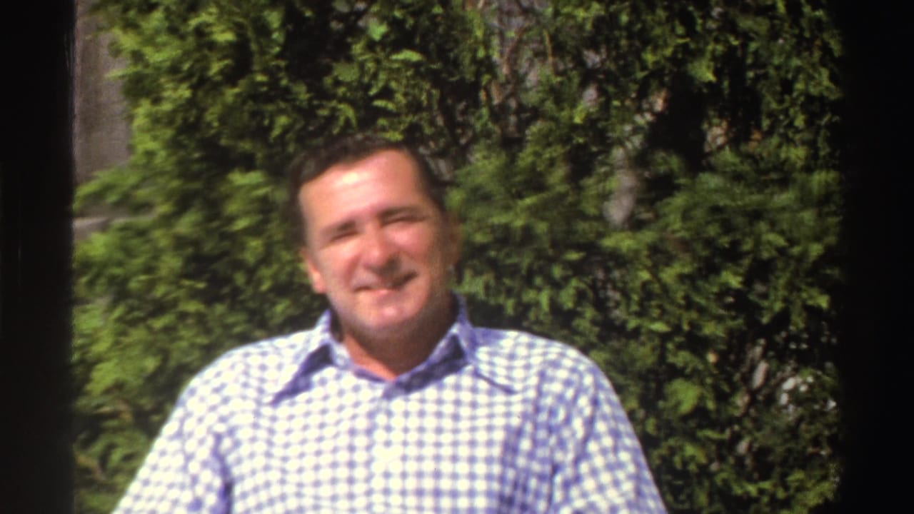 In 1969 Michigan home-movie footage, an adult man in a blue and white checkered shirt faces the camera in a sunny garden.
