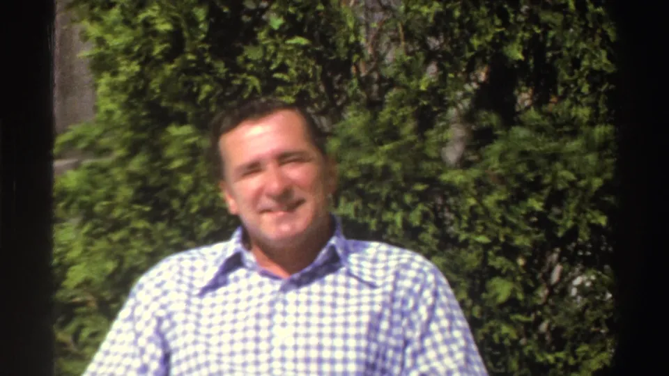 In 1969 Michigan home-movie footage, an adult man in a blue and white checkered shirt faces the camera in a sunny garden.