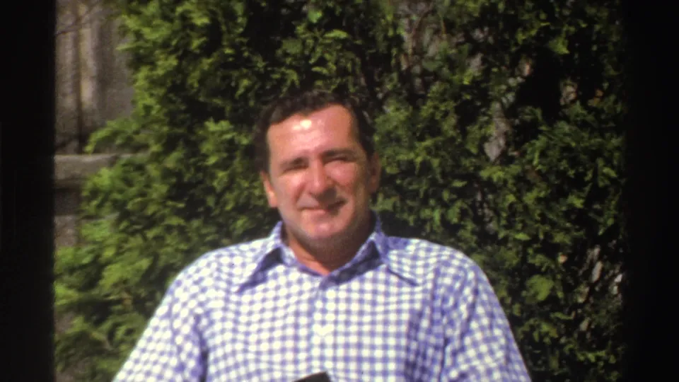 A smiling adult man stands outdoors in bright light, with dense garden foliage behind him in this Michigan 1969 8mm frame.