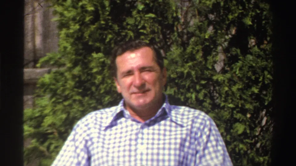 An adult man in a checkered shirt faces the camera, with shrubs and a partial masonry surface visible behind him.