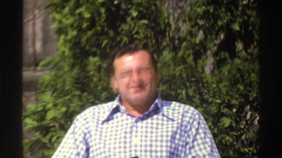 A soft-focused 8mm frame shows an adult man in a checkered shirt standing in bright outdoor light before green foliage.