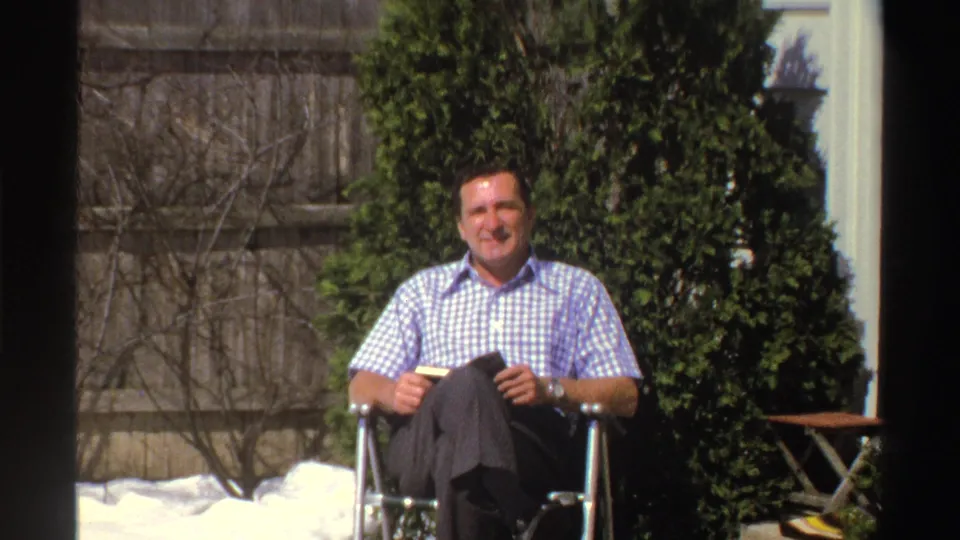 A man in a blue and white checkered shirt sits outdoors in a snowy Michigan yard, holding a small object near a wooden fence and evergreens.