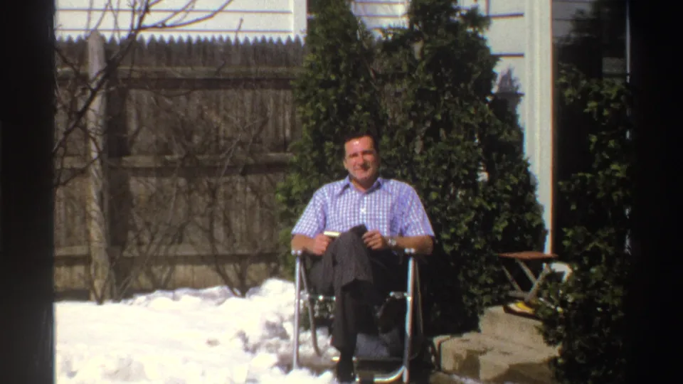 Wider 8mm frame shows a seated man in a snowy Michigan backyard, framed by a wooden fence, bare branches, evergreens, and house siding.