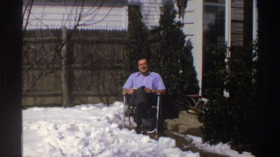 A distant 1969 Michigan home-movie frame shows a man seated in a folding chair beside snow, evergreen shrubs, a wooden fence, and a house exterior.