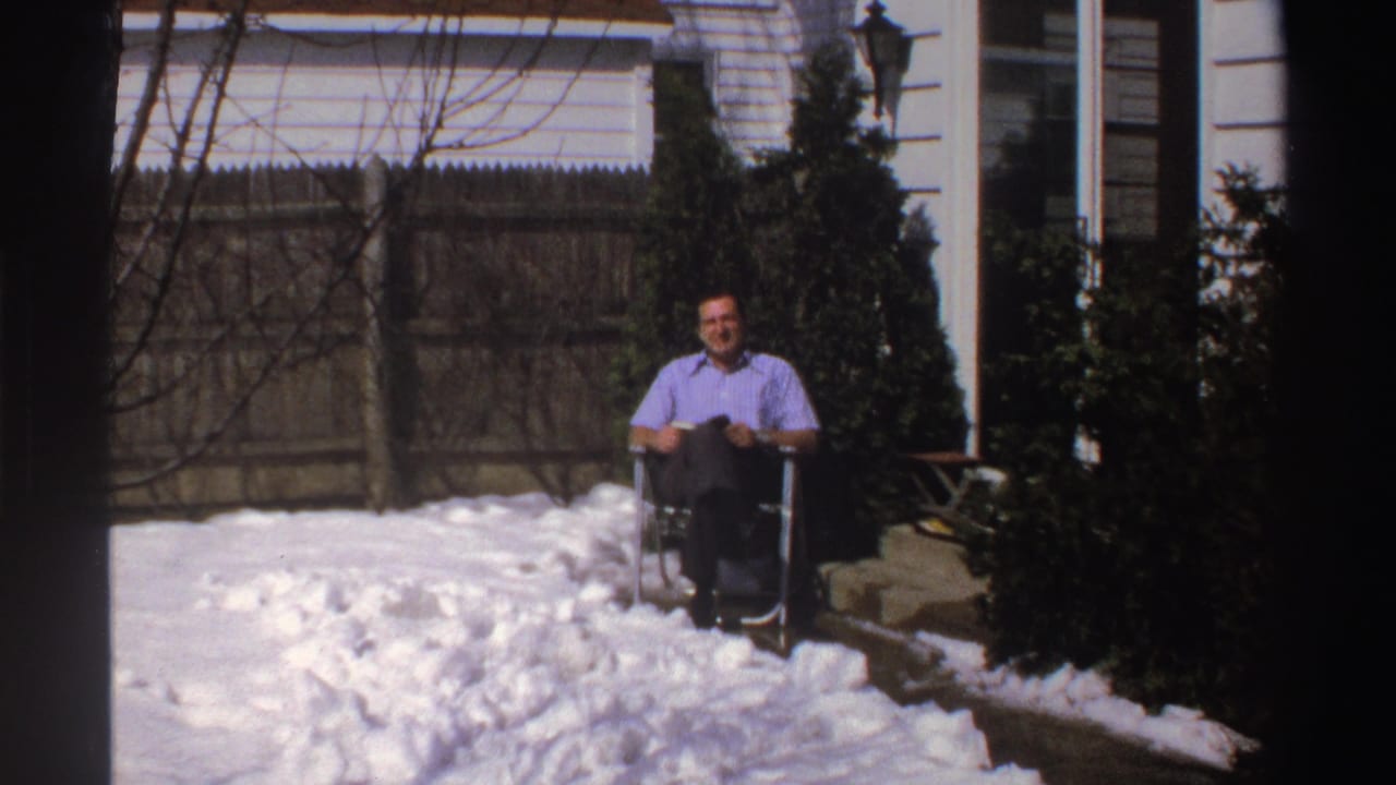 In a 1969 Michigan home-movie frame, a man sits smiling in a folding chair amid deep backyard snow.