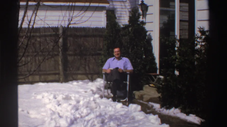 In a 1969 Michigan home-movie frame, a man sits smiling in a folding chair amid deep backyard snow.
