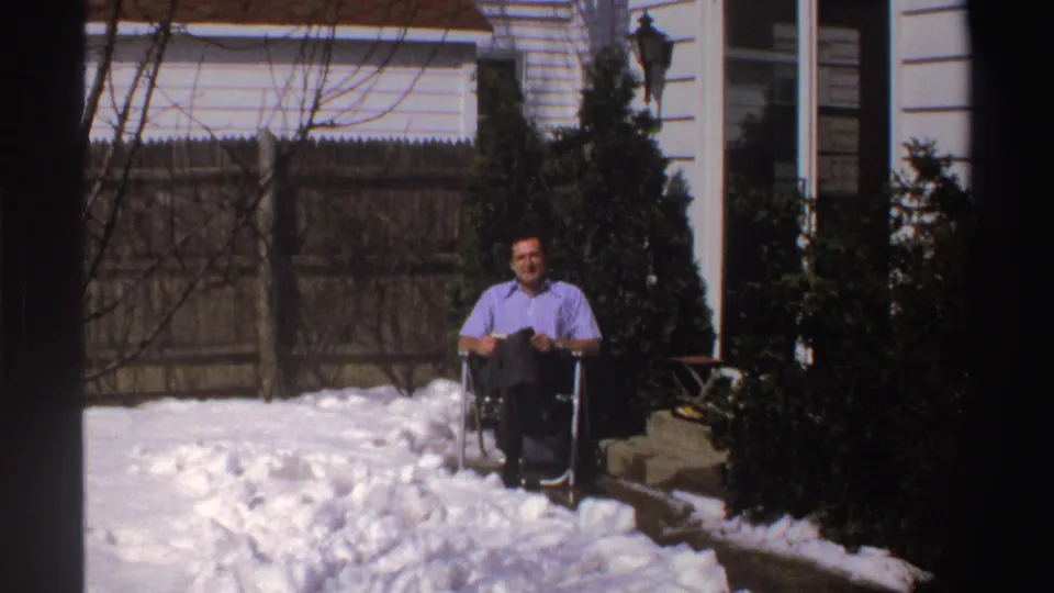 The man remains seated in the snowy Michigan backyard, facing the camera in soft 8mm light.