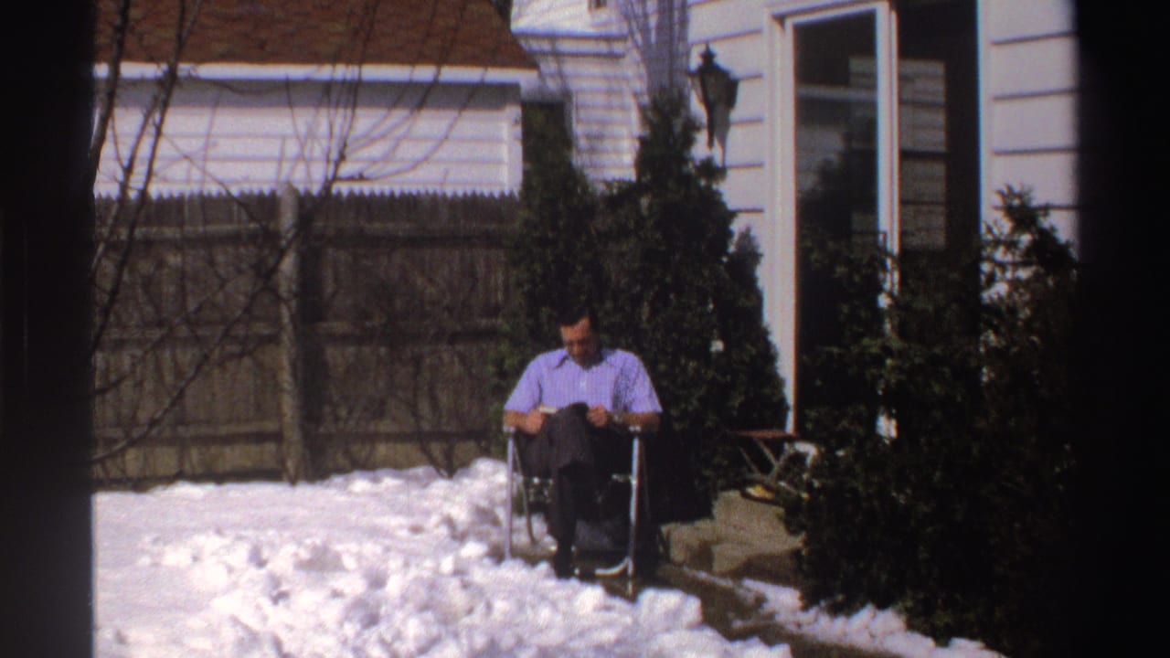 Man seated in a folding chair reading in a snowy backyard beside shrubs and a white house.