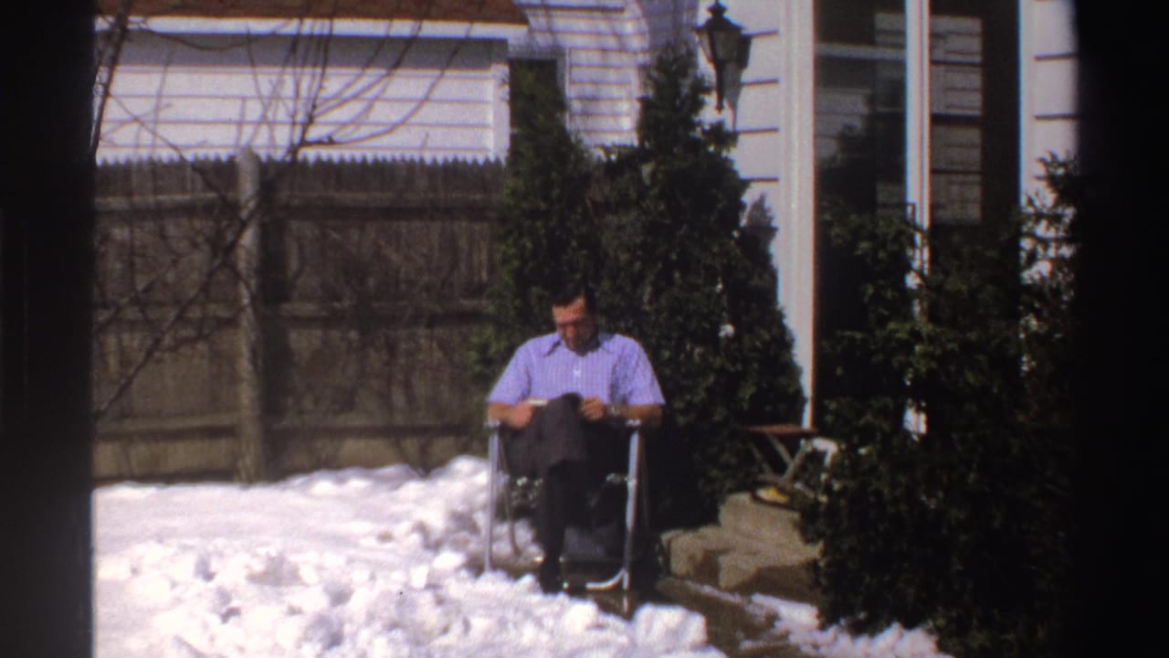 The man remains seated and reading in the snowy Michigan backyard, framed by a wood fence, shrubs, house siding, and a glass door.